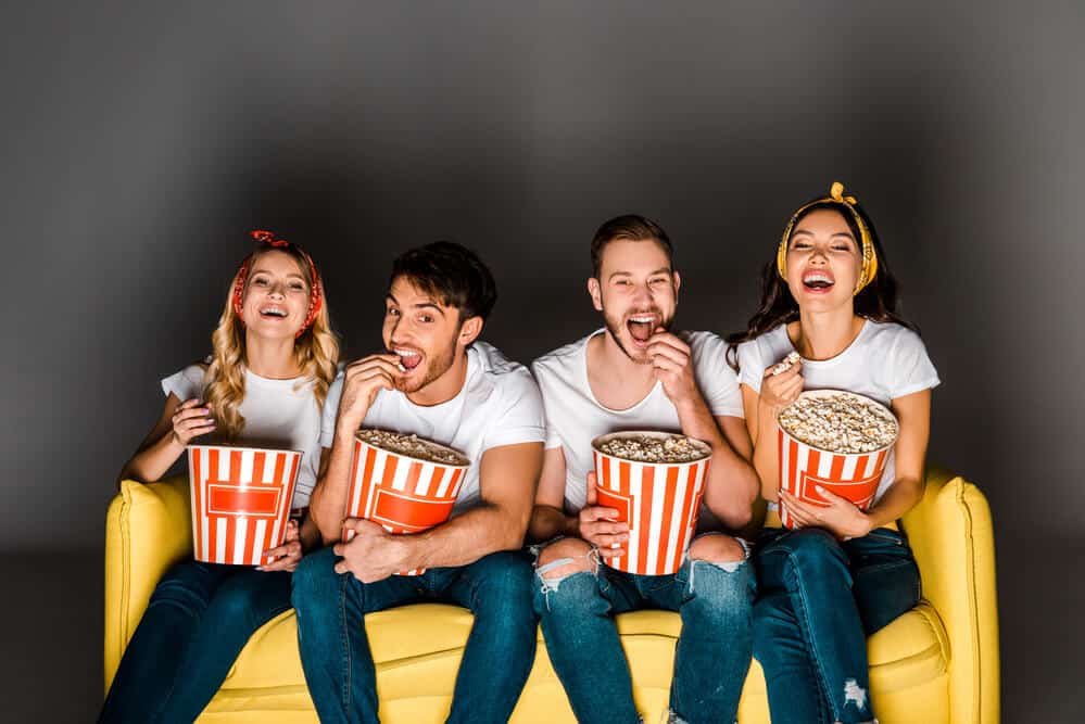 the positive effects from watching tv are laughing and bonding with friends - actual image of four friends laughing on a yellow couch watching tv and eating popcorn